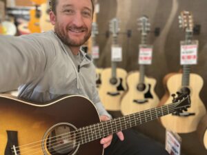 This is a photo of Brad Johnson, the writer and owner of Song Production Pros playing a Taylor American Dream AD17e. He is wearing a white sweatshirt and hat in a guitar shop room.