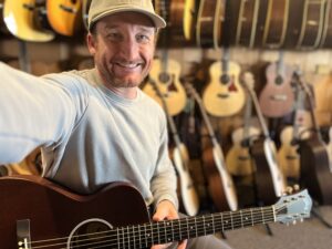 This is a photo of Brad Johnson, the writer and owner of Song Production Pros playing a Guild M-20. He is wearing a blue sweatshirt and green hat in a guitar shop room.