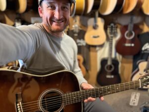 This is a photo of Brad Johnson, the writer and owner of Song Production Pros playing a Martin D-35. He is wearing a blue sweatshirt and green hat in a guitar shop room.