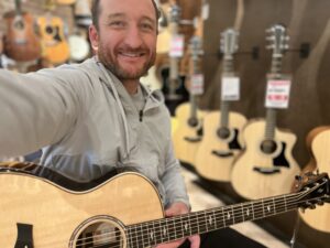 This photo shows the founder and writer of Song Production Pros playing a Taylor 814ce acoustic guitar. He is wearing a grey jacket and is in a guitar store.